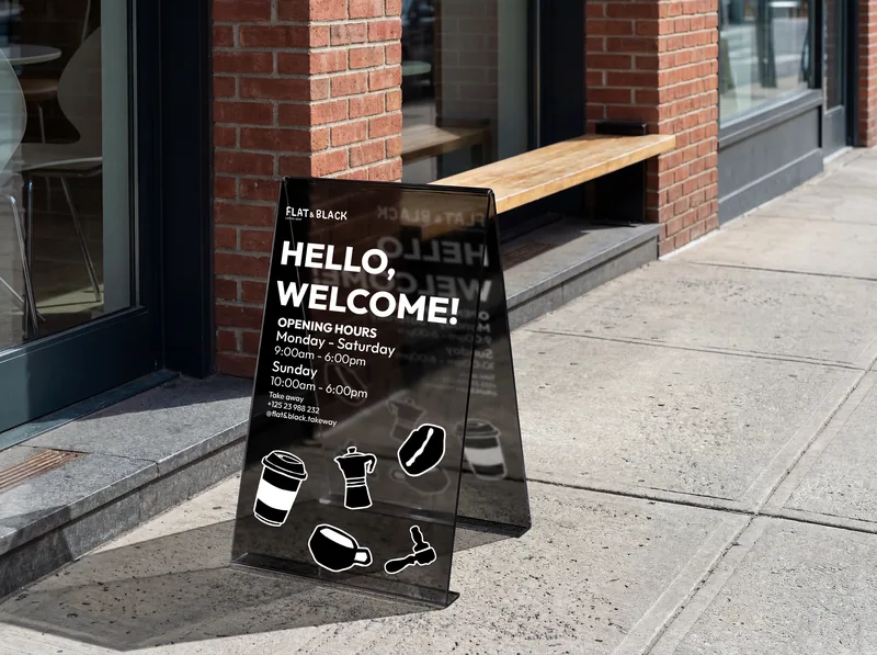 A black A-board on the footpath outside a brick shopfront: Hello, welcome, the opening hours, and the pictograms tumbling down the panel.